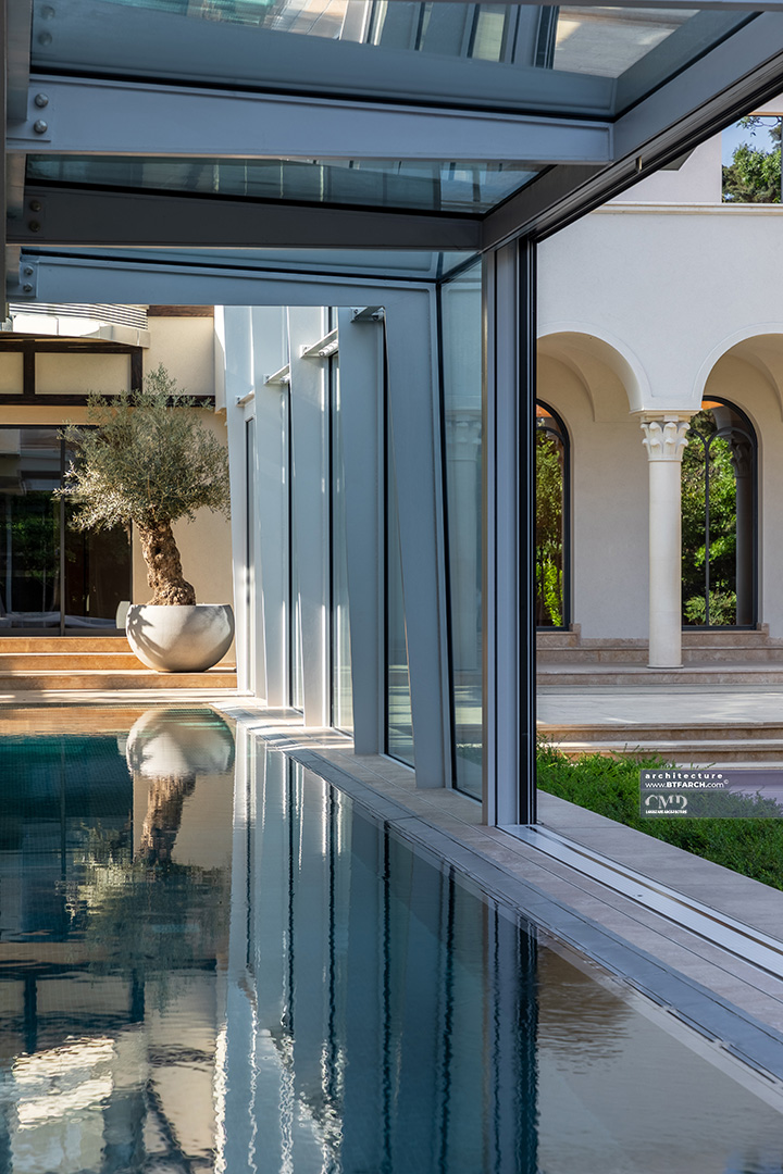 Solitary olive tree in a large planter next to a glass-enclosed indoor pool and Mediterranean architecture